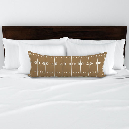 Stadium Stripes gold football lumbar pillow styled on a bed — gold lumbar pillow cover on white bedding with dark wood headboard