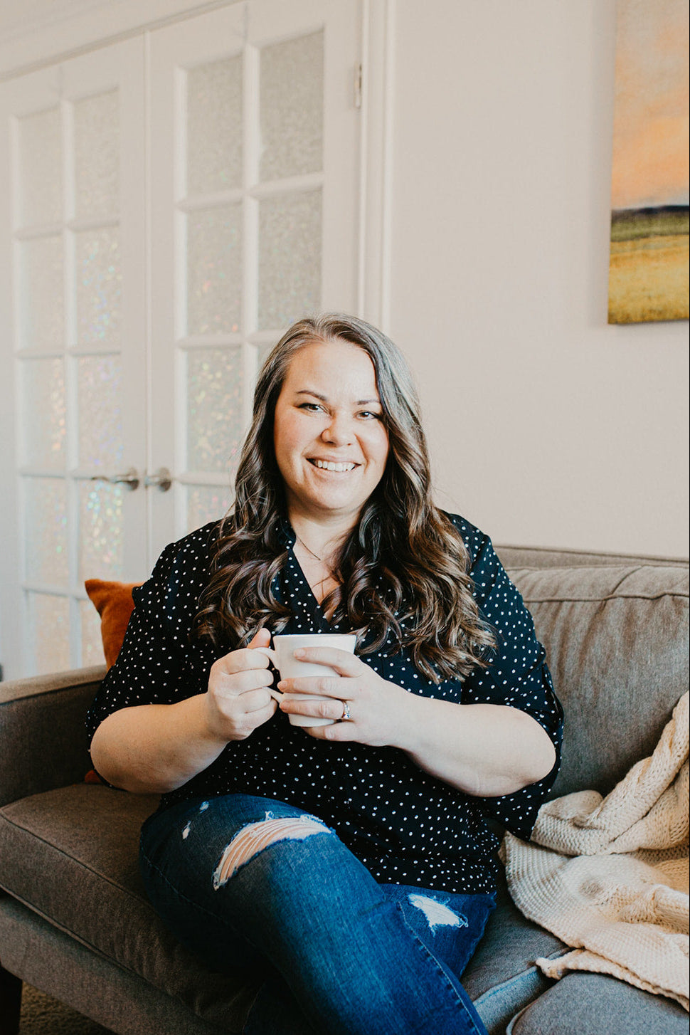 A smiling woman with long brown hair sits on a gray couch holding a white mug, wearing a black short-sleeve top with white polka dots and blue jeans with a ripped knee detail.